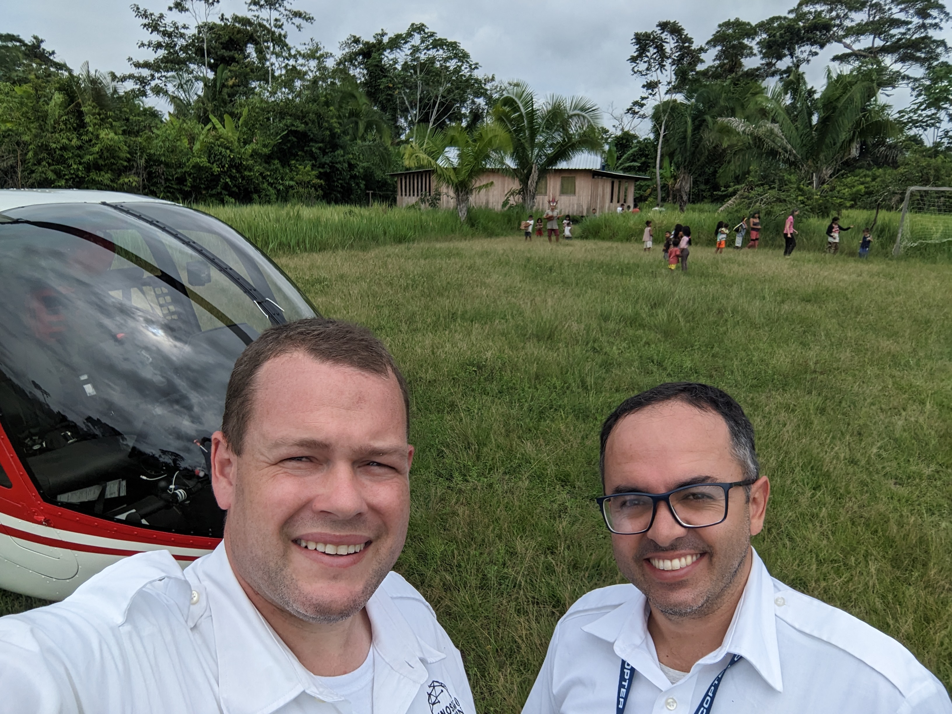 Two male helicopter pilots stand in front of an R66 helicopter and a group of children playing in Brazil.