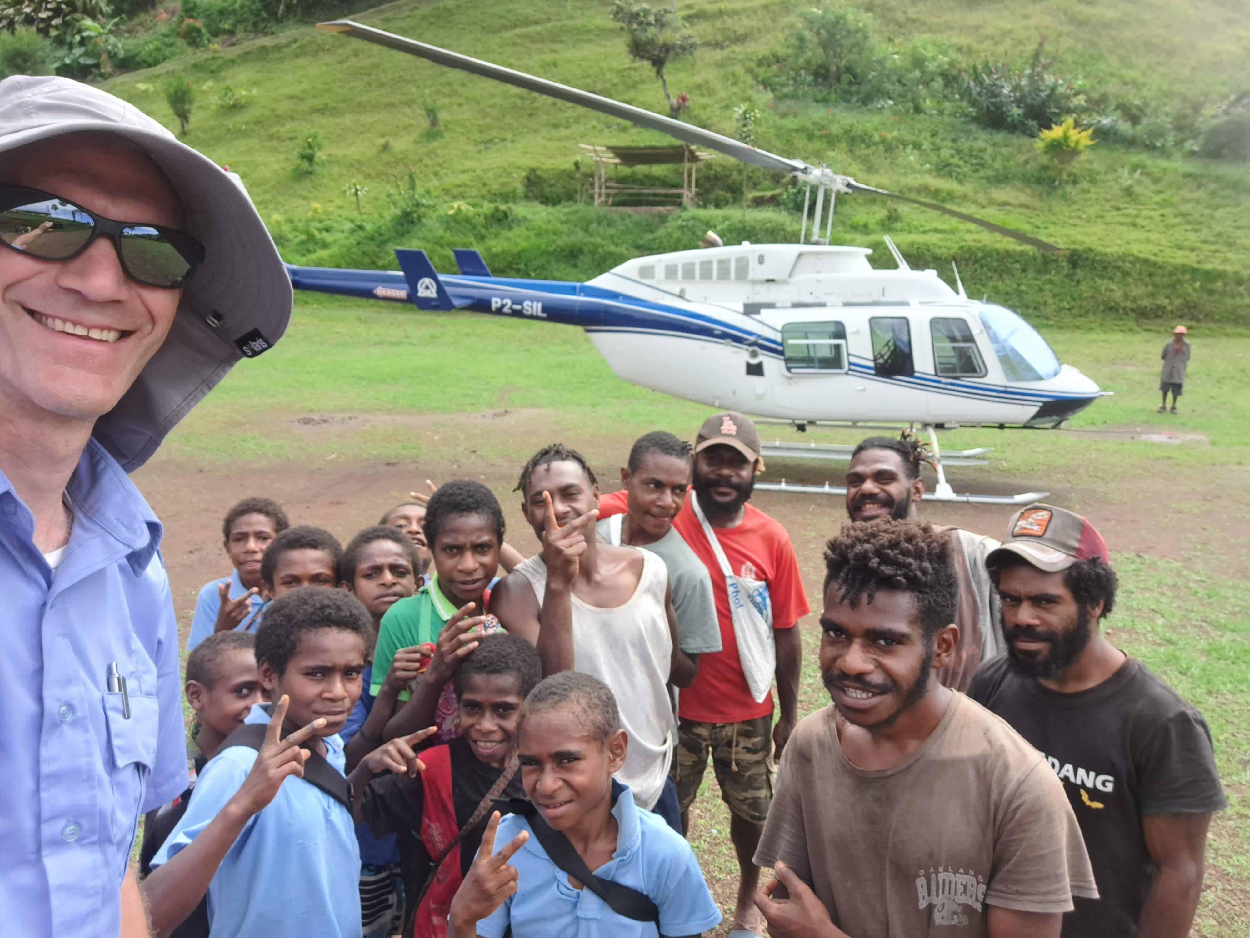 A helicopter pilot stands besides a group of Papua New Guinean boys and ben with a helicopter in the background.
