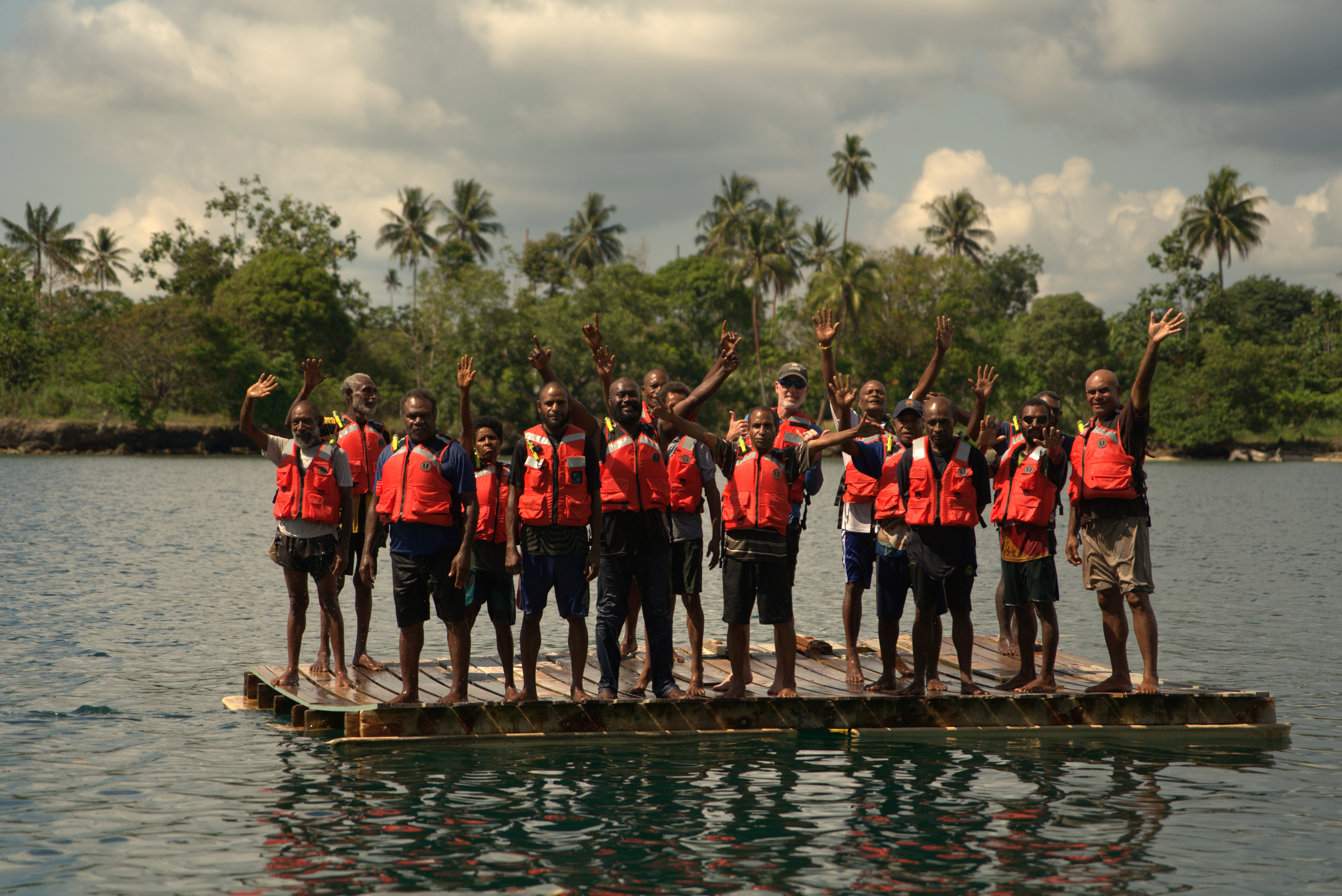 A group of Papua New Guineans with life jackets stand with an American on a floating platform in the water while waving at the camera